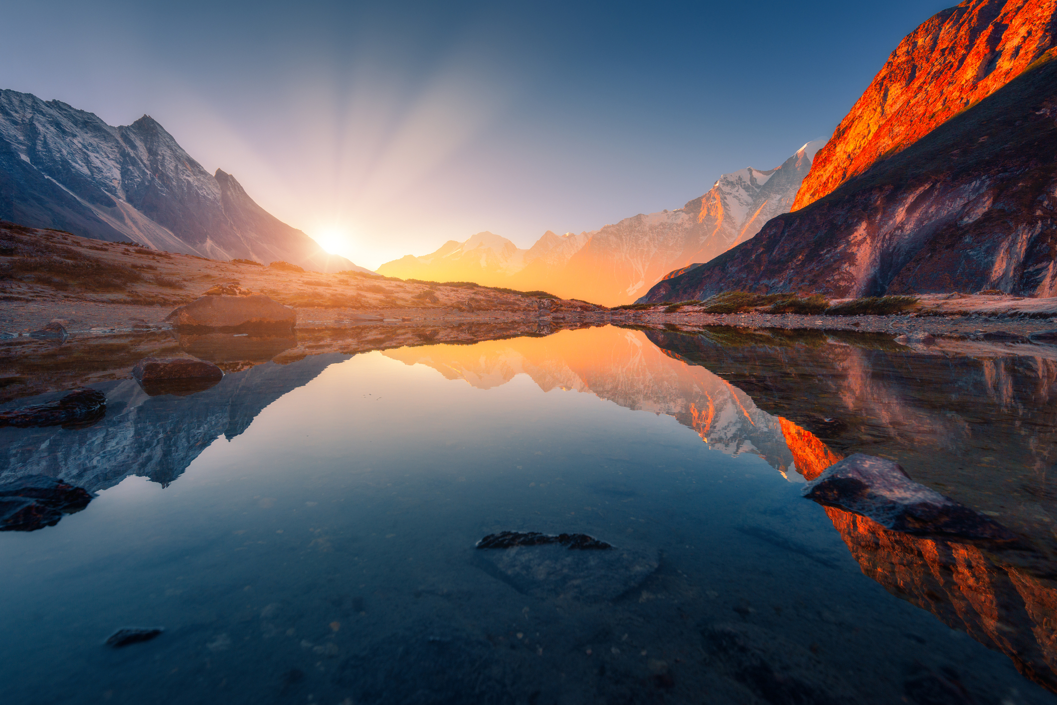 Beautiful landscape with high mountains with illuminated peaks, stones in mountain lake, reflection, blue sky and yellow sunlight in sunrise. Nepal. Amazing scene with Himalayan mountains. Himalayas